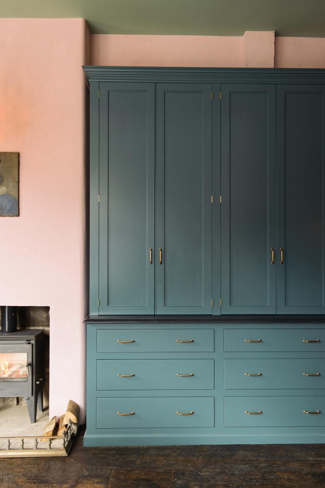 The playful pastel yet classic kitchen features a large Clerkenwell blue full-height pantry cupboard with a black granite worktop and a pink chimney with a small iron wood stove. Decorated with chopped firewood and vintage ceramics, this kitchen has both functionality and personality.