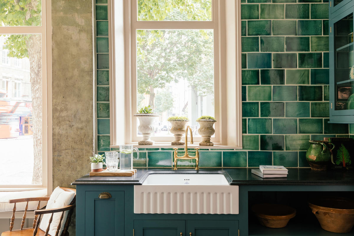 The playful pastel yet classic kitchen features a Clerkenwell blue sink run with a black granite worktop, emerald green splashback tiles, a single fluted Belfast sink, and an aged brass Ionian tap. Decorated with vintage ceramics, furniture and potted plants, this kitchen has both functionality and personality.