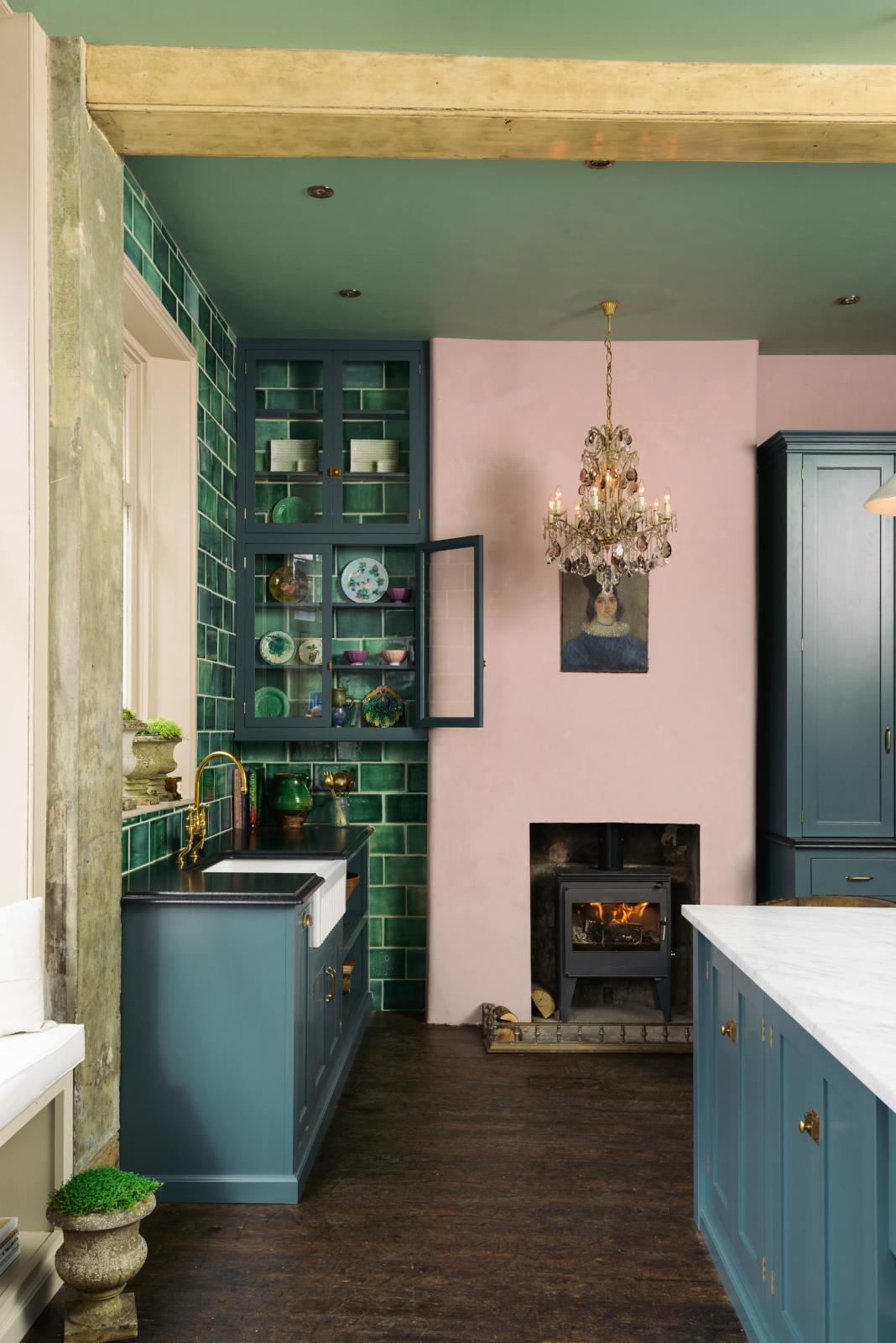 The kitchen features a blend of playful pastels and classic design, with a Carrara marble island, wall cupboards, and a Clerkenwell blue sink. It includes a full-height pantry with a black granite worktop, emerald green splashback tiles, and a pink chimney with a small wood stove. Decorated with firewood, vintage ceramics, an oil portrait, and a crystal chandelier, this space balances functionality and style.