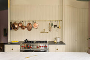 A beige cooker run with a black granite worktop, a silver gas cooker, an aged brass hanging rail, an aged brass pot filler and a long kitchen island with a Carrara marble worktop. Lovingly decorated with hanging copper pots and pans, fresh lemons, garlic, chilli and sage, this space balances functionality and style.