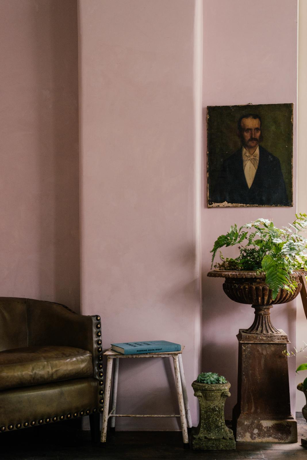 A serene corner of a pastel-colored classic kitchen is beautifully adorned with vintage furniture, including an armchair, a stool, garden ornaments, vibrant plants, and an oil painting.