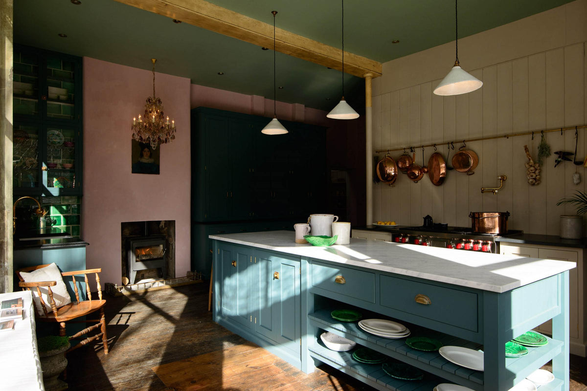 A dramatic image inside the deVOL St John's Square, London showroom. The spacious room shows a playful pastel yet classic kitchen features a mushroom-coloured cooker run, aged brass hanging rails, and a pot filler. A long kitchen island with a breakfast nook and deVOL bum stools adds charm. Finally, a Clerkenwell blue sink run with a black granite worktop, and emerald green splashback tiles. Decorated with copper pots, vintage ceramics, and fresh produce, this kitchen has both functionality and personality.