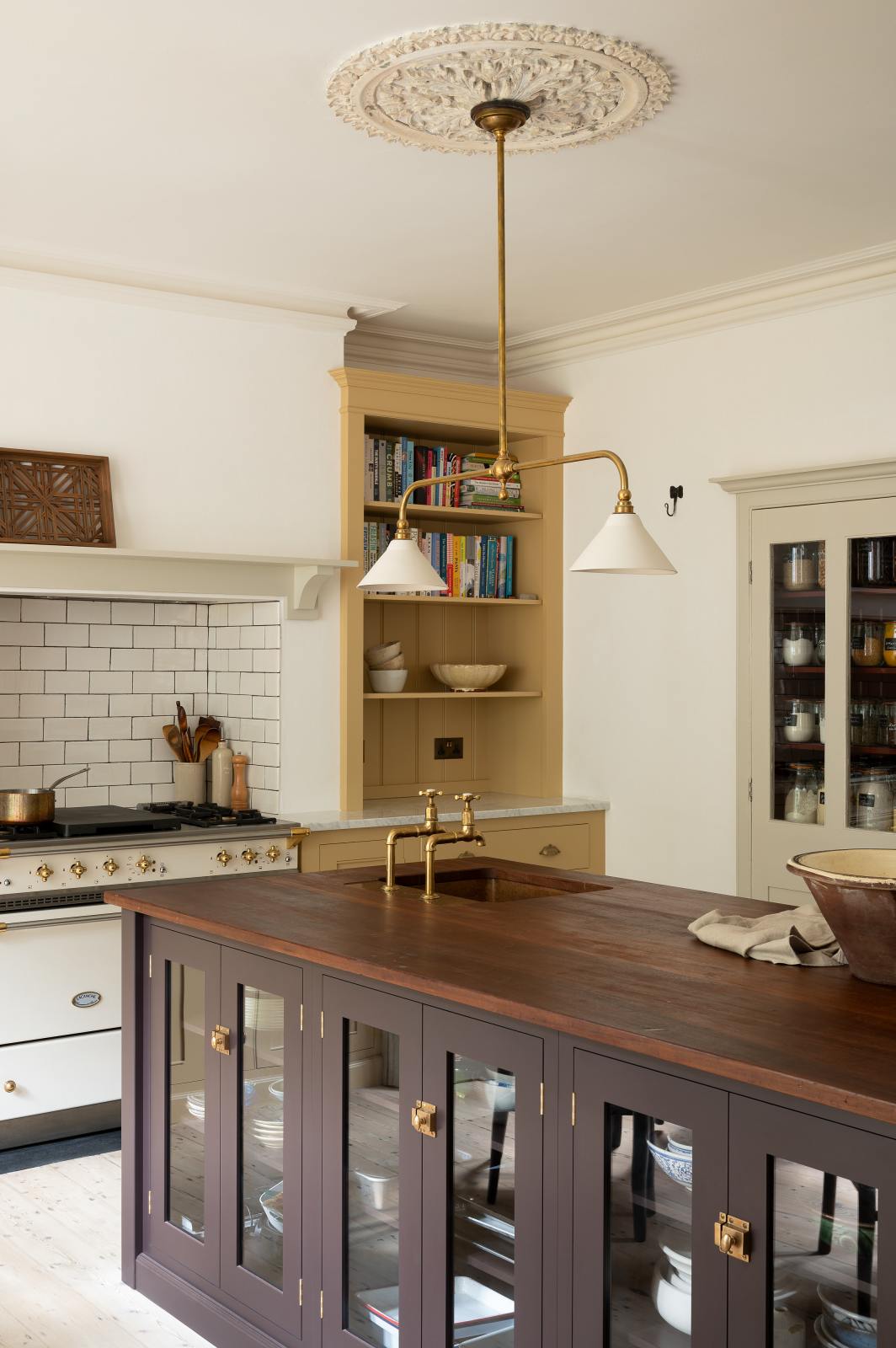 An oblique view of a lovely and simple blush yellow, classic English kitchen features a cooker run with Carrara marble worktop, two open countertop cupboards, a white gas stove inside a chimney breast, porcelain splashback, a purple-brown kitchen island with a solid oak worktop and aged brass Mayan taps. A deVOL gaselier light and a collection of muted ceramics and tall wildflowers create an elegant and inviting atmosphere in the room.