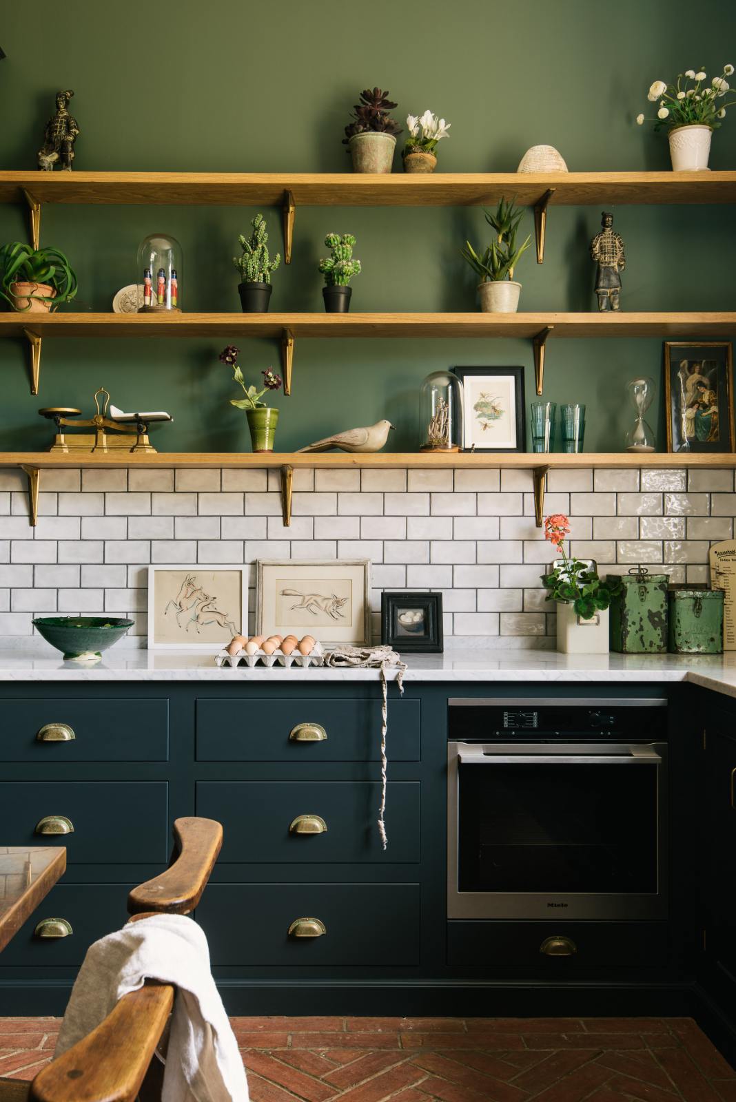 The deep blue base cupboards contain an under-counter oven and are topped with Carrara marble. A lovely white metro tile splashback extends up the wall to meet the first three wooden shelves, each supported by beautiful handmade aged brass brackets. The walls are painted a muted dark green, and the shelves display an array of trinkets, plants, and artwork.