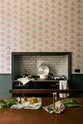 The image features a close-up view of an aged copper tabletop in the foreground. In the background, there is a chimney breast with a black Aga stove situated in the alcove. The chimney breast is adorned with whimsical wallpaper that has a pale cream base colour, decorated with illustrations of light pink jellies and cakes.