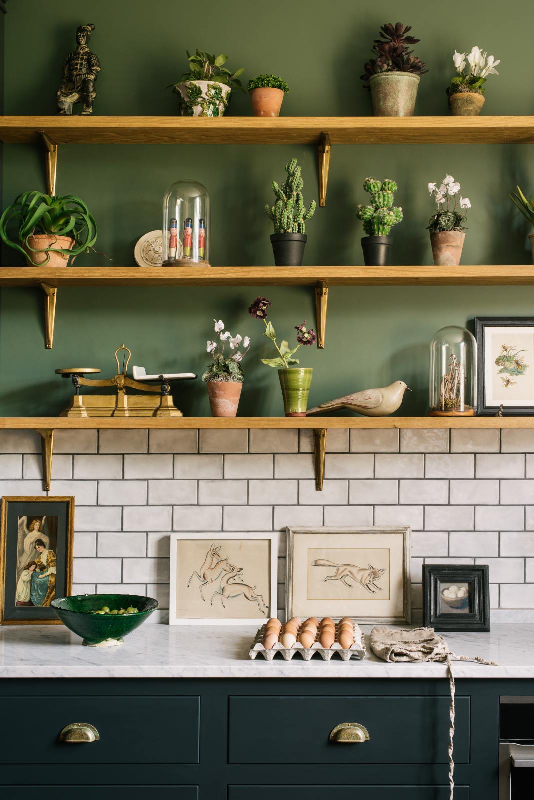 The deep blue base cupboards contain an under-counter oven and are topped with Carrara marble. A lovely white metro tile splashback extends up the wall to meet the first three wooden shelves, each supported by beautiful handmade aged brass brackets. The walls are painted a muted dark green, and the shelves display an array of trinkets, plants, and artwork.