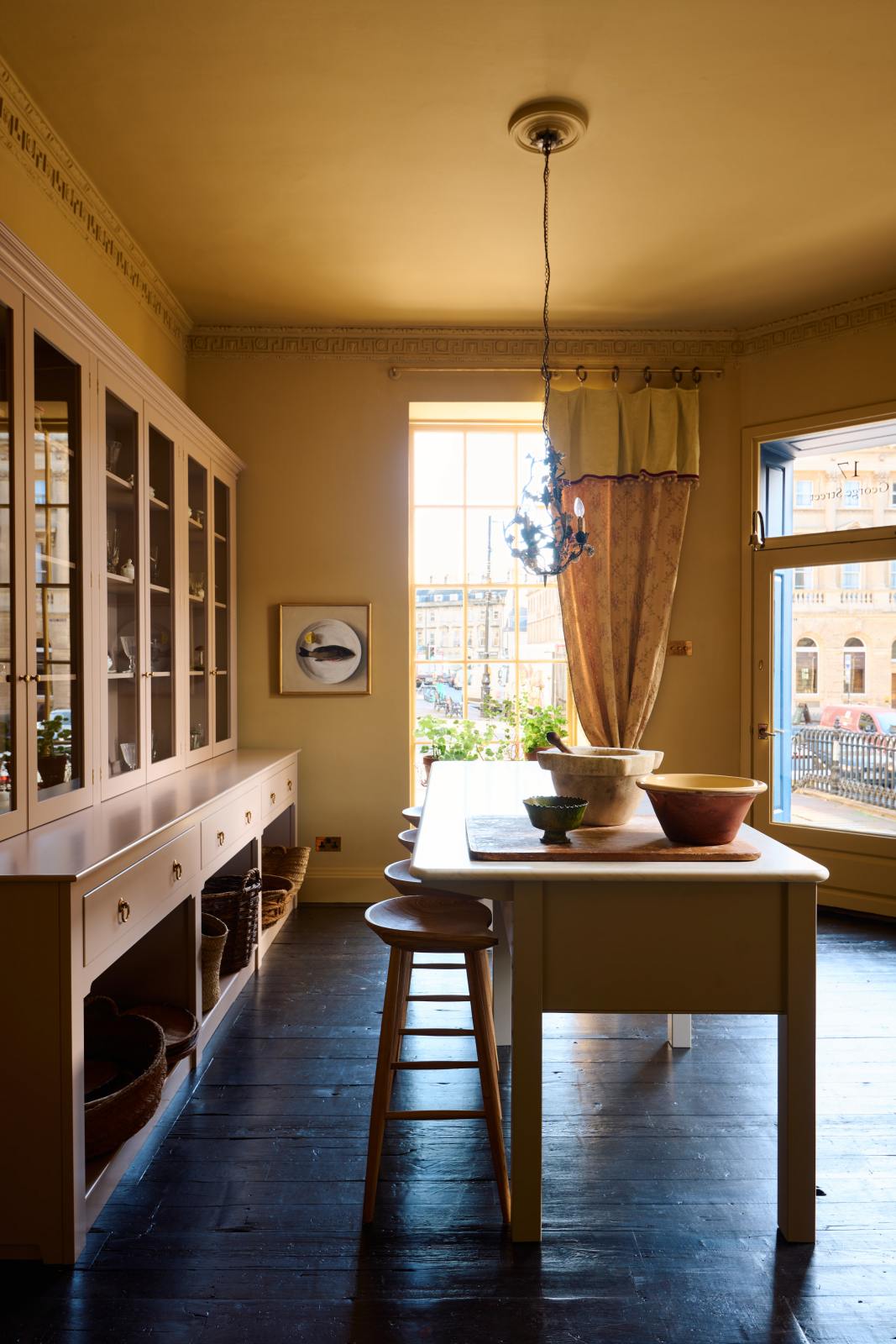 A warm yellow room with a large Princelet pink dresser showcasing glazed cupboards filled with delicate ceramics, papier-mâché mushrooms, and wooden baskets along the bottom, giving the space a relaxed, living-room feel. The space includes a wide prep table with an open nook for deVOL bum stools. There are large ceramic bowls, a metal vines-and-flowers chandelier, and a golden-framed painting of a fish and a lemon.