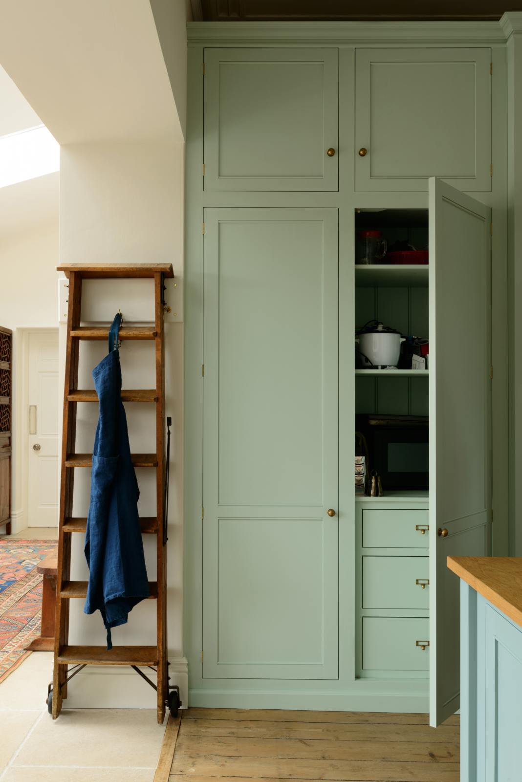 A corner of a humble and calming turquoise, classic English kitchen featuring a large full-height pantry cupboards with top cabinets, and a wooden ladder. Adorned with navy blue apron, a small rice cooker, and a silver cheese grater, it creates a warm and simple home kitchen.