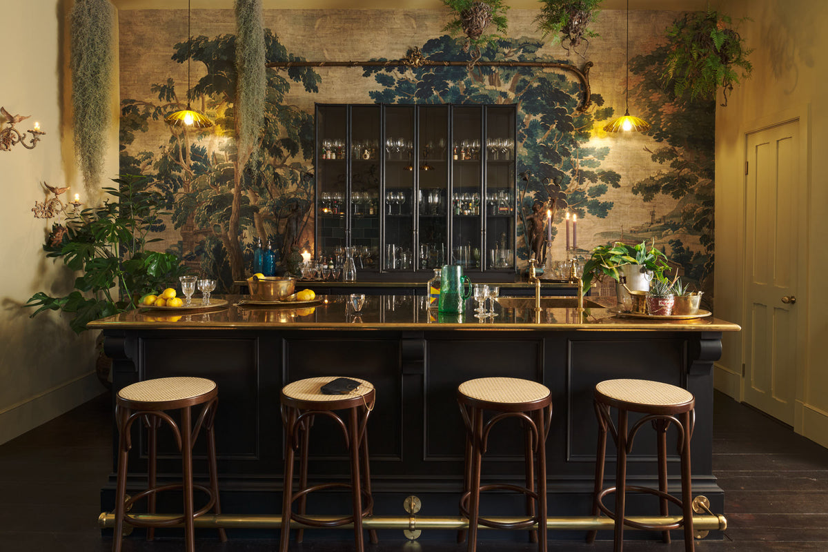 A chic and glamorous Bobbin Black Haberdasher’s bar featuring a kitchen island with a polished brass worktop, a single polished brass sink, aged brass Mayan taps, a brass foot rail, glazed base cabinet with internal drawers, tall countertop cupboards, and woven top bentwood café stools. Set against an illustrated foliage wallpaper and adorned with hanging plants, wine glasses, and vintage decanters, it creates a beautiful and intimate personal bar.