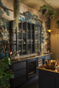 An oblique view of a chic and glamorous Bobbin Black Haberdasher’s bar featuring a kitchen island with a polished brass worktop, glazed base cabinet with internal drawers, and tall countertop cupboards. Set against an illustrated foliage wallpaper and adorned with hanging plants, wine glasses, and vintage decanters, it creates a beautiful and intimate personal bar.