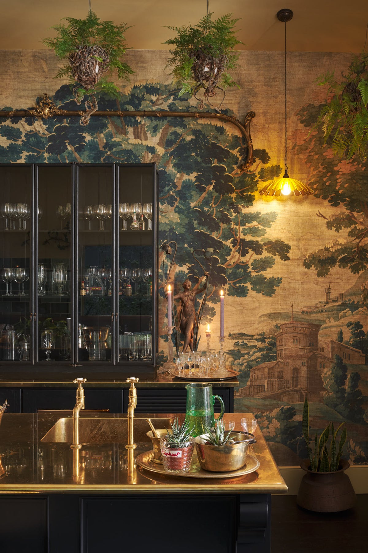 A quiet corner of a chic and glamorous Bobbin Black Haberdasher’s bar featuring a kitchen island with a polished brass worktop, a single polished brass sink, aged brass Mayan taps, glazed base cabinet with internal drawers, and tall countertop cupboards. Set against an illustrated foliage wallpaper and adorned with hanging plants, wine glasses, and vintage decanters, it creates a beautiful and intimate personal bar.