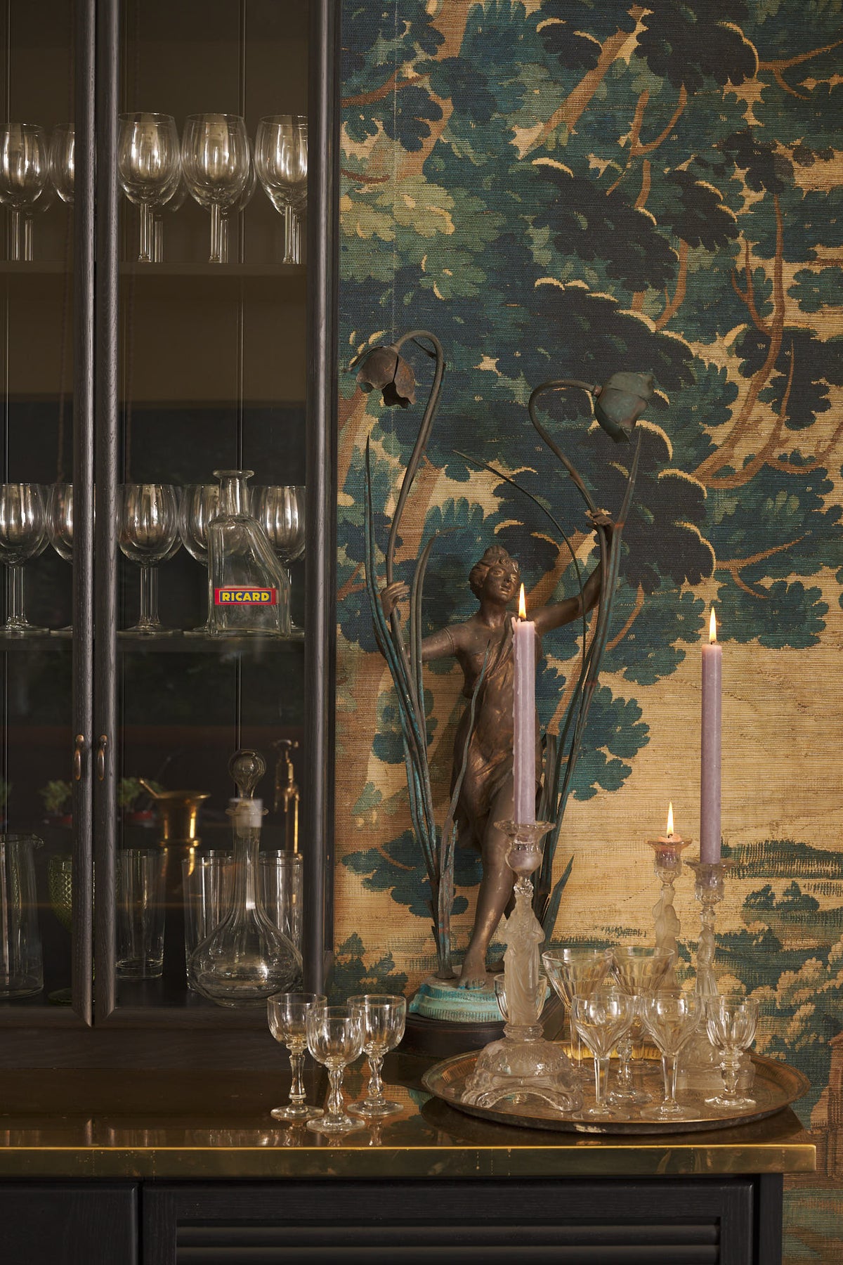 A close-up of a chic and glamorous Bobbin Black Haberdasher’s bar featuring glazed base cabinets with internal drawers, and tall countertop cupboards. Set against an illustrated foliage wallpaper and adorned with wine glasses, an antique brass figurine, crystal handle holders, and vintage decanters, it creates a beautiful and intimate personal bar.