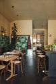 A chic and glamorous Bobbin Black Haberdasher’s bar featuring a kitchen island with a polished brass worktop, a brass foot rail, vineyard tables, bentwood café chairs, woven top bentwood café stools, petite amber frilly glass pendant lights, and a large display of Emerald green London splashback tiles. Adorned with wine glasses, plants, crystal handle holders, trays, and vintage decanters, it creates a beautiful and intimate personal bar.