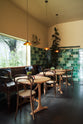 An oblique view of a chic and glamorous Bobbin Black Haberdasher’s bar featuring vineyard tables, bentwood café chairs, petite amber frilly glass pendant lights, and a large display of Emerald green London splashback tiles. Adorned with a long antique couch, plants, vintage watering cans, and deVOL candles, it creates a beautiful and intimate personal bar.