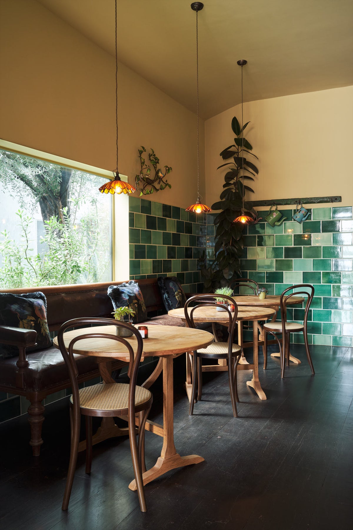 An oblique view of a chic and glamorous Bobbin Black Haberdasher’s bar featuring vineyard tables, bentwood café chairs, petite amber frilly glass pendant lights, and a large display of Emerald green London splashback tiles. Adorned with a long antique couch, plants, vintage watering cans, and deVOL candles, it creates a beautiful and intimate personal bar.