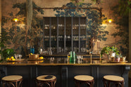 A chic and glamorous Bobbin Black Haberdasher’s bar featuring a kitchen island with a polished brass worktop, a single polished brass sink, aged brass Mayan taps, a brass foot rail, glazed base cabinet with internal drawers, tall countertop cupboards, and woven top bentwood café stools. Set against an illustrated foliage wallpaper and adorned with hanging plants, wine glasses, and vintage decanters, it creates a beautiful and intimate personal bar.