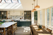 An elegant and spacious Haberdasher’s kitchen features a large cooker run, a kitchen island with a double Belfast sink, an aged brass Ionian tap, an Arabescato marble worktop, splashback, and glazed cupboards. Adorned with vintage furniture, artisan ceramics, glass flower pendant lights, lemons, and limes, this kitchen creates a classic social space with functional storage.