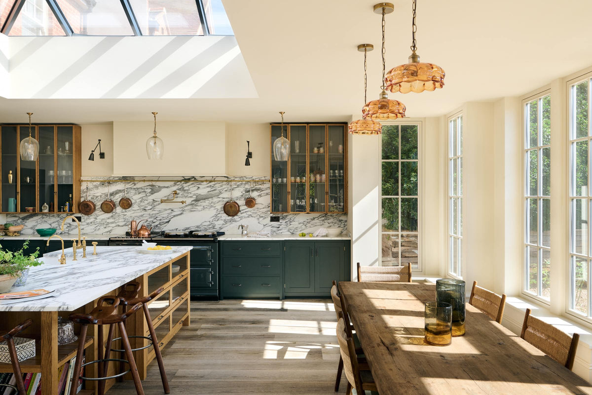 An elegant and spacious Haberdasher’s kitchen features a large cooker run, a kitchen island with a double Belfast sink, an aged brass Ionian tap, an Arabescato marble worktop, splashback, and glazed cupboards. Adorned with vintage furniture, artisan ceramics, glass flower pendant lights, lemons, and limes, this kitchen creates a classic social space with functional storage.