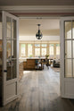 Past white French doors is an elegant and spacious Haberdasher’s kitchen features a large cooker run, a kitchen island with a double Belfast sink, an aged brass Ionian tap, an Arabescato marble worktop, splashback, and glazed cupboards. Adorned with vintage furniture, artisan ceramics, glass flower pendant lights, and a sunflower arrangement, this kitchen creates a classic social space with functional storage.