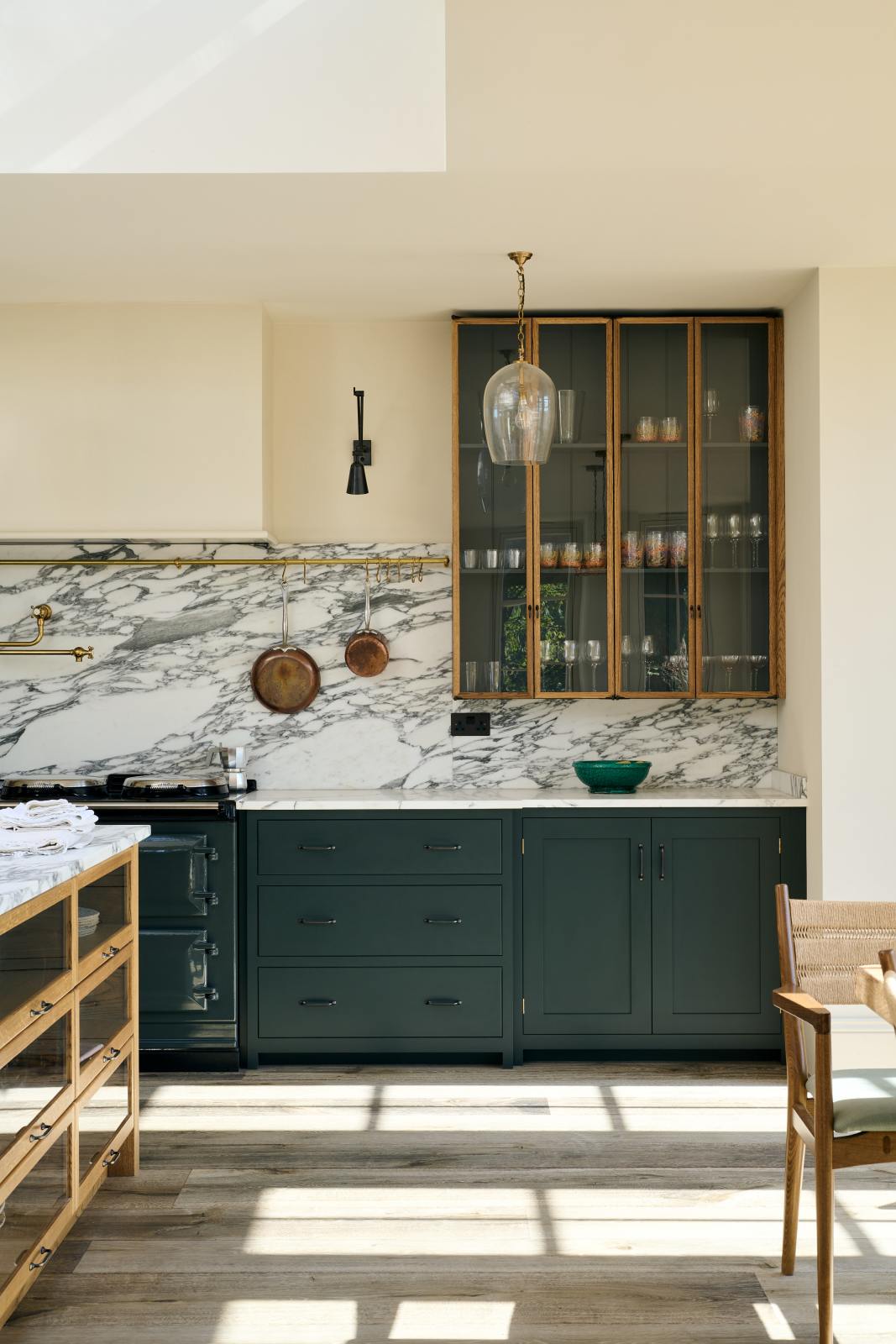 A quiet corner of an elegant and spacious Haberdasher’s kitchen features a large cooker run, a kitchen island with an Arabescato marble worktop, a splashback, and glazed cupboards. Adorned with vintage furniture, artisan ceramics, glass pendant lights, copper pans, and a bowl of lemons, this kitchen creates a classic social space with functional storage.