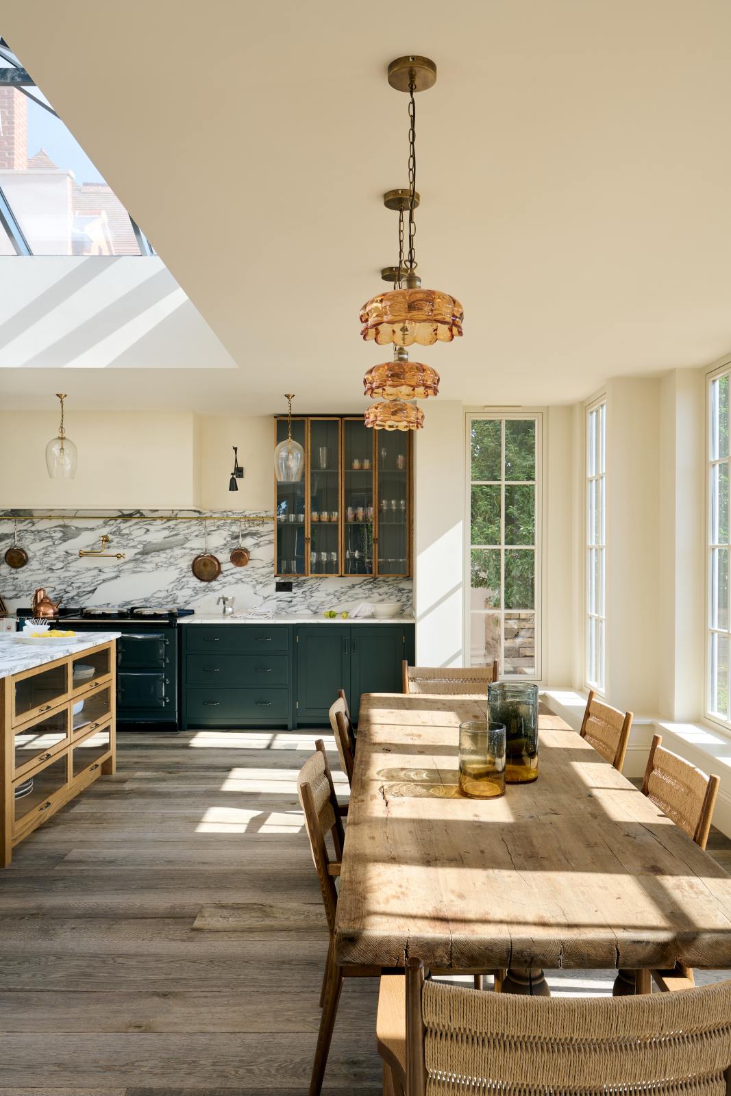 An elegant and spacious Haberdasher’s kitchen features a large cooker run, a kitchen island with an Arabescato marble worktop, splashback, and glazed cupboards. Adorned with vintage furniture, artisan ceramics, glass flower pendant lights, and limes, this kitchen creates a classic social space with functional storage.