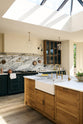 An oblique view of an elegant and spacious Haberdasher’s kitchen features a large cooker run, a kitchen island with a double Belfast sink, an aged brass Ionian tap, an Arabescato marble worktop, splashback, and glazed cupboards. Adorned with vintage furniture, artisan ceramics, glass pendant lights, lemons, and a skylight, this kitchen creates a classic social space with functional storage.