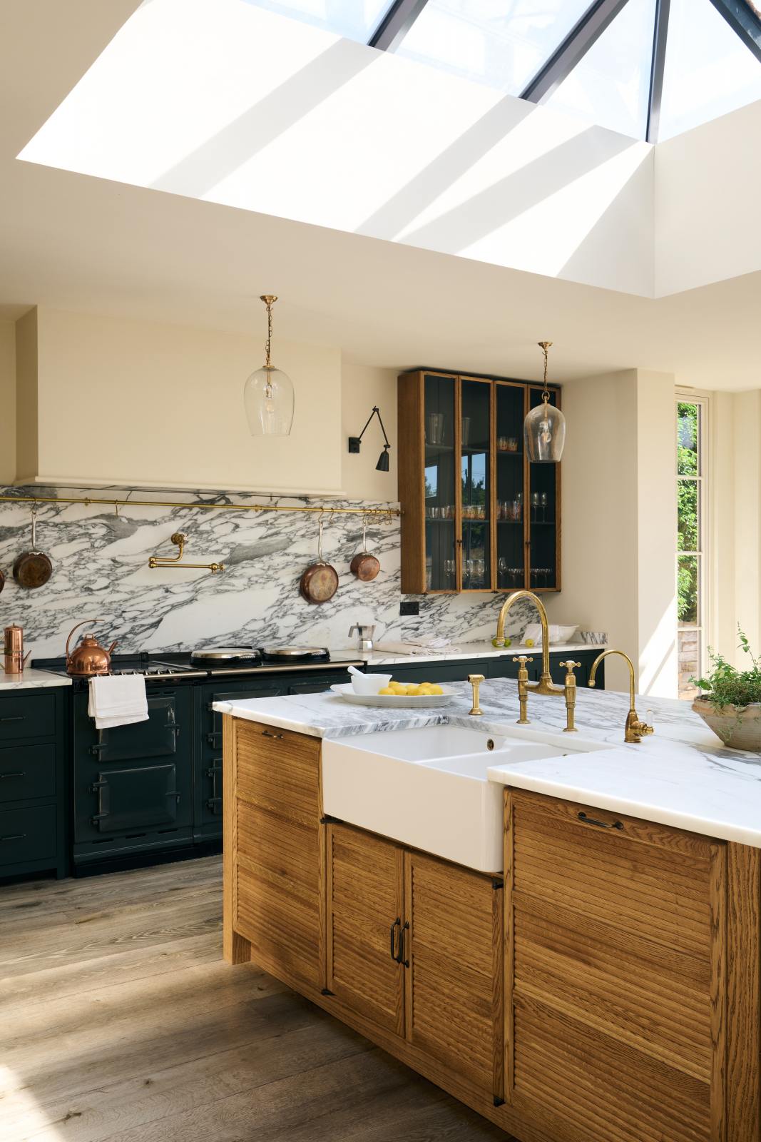 An oblique view of an elegant and spacious Haberdasher’s kitchen features a large cooker run, a kitchen island with a double Belfast sink, an aged brass Ionian tap, an Arabescato marble worktop, splashback, and glazed cupboards. Adorned with vintage furniture, artisan ceramics, glass pendant lights, lemons, and a skylight, this kitchen creates a classic social space with functional storage.
