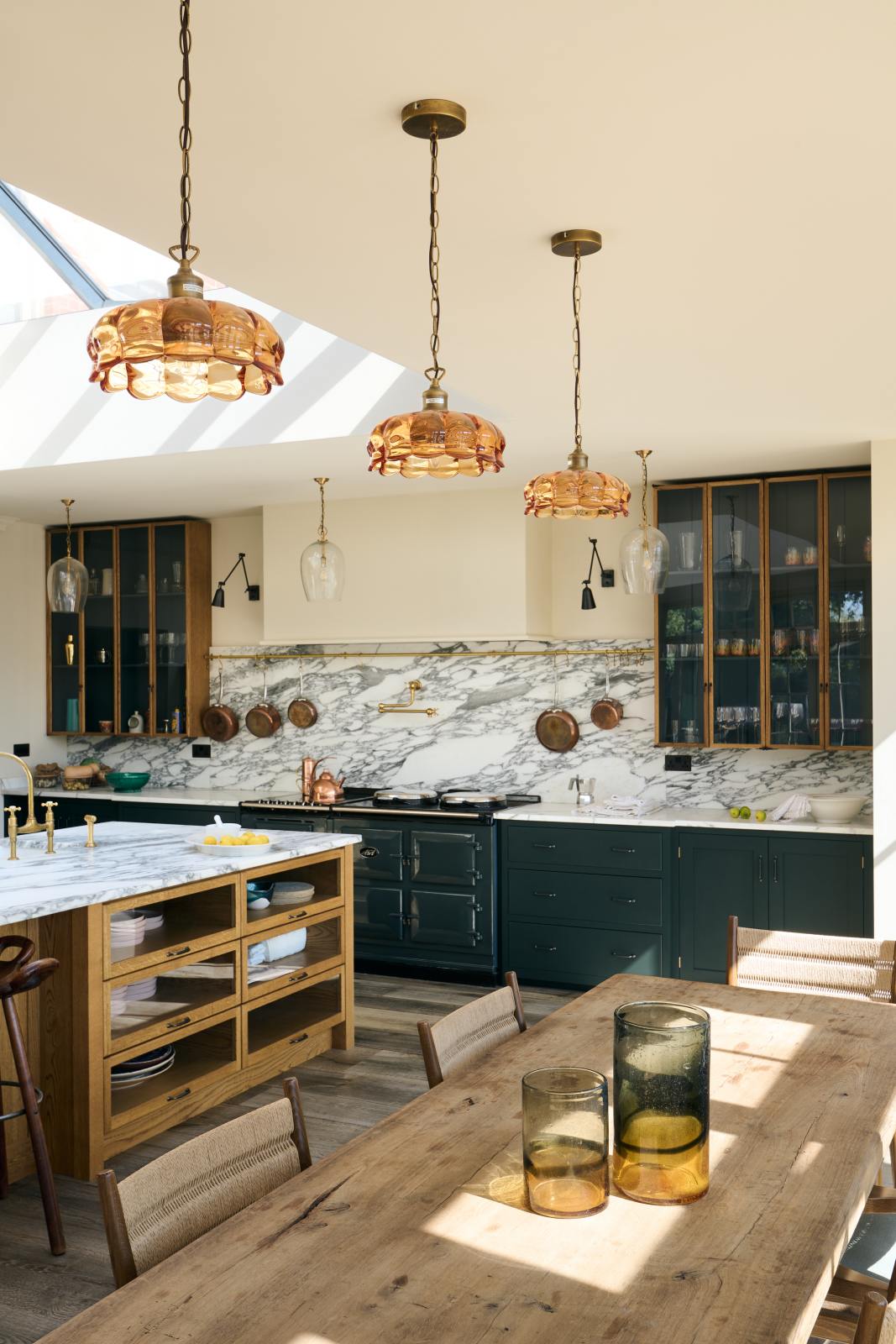 An oblique view of an elegant and spacious Haberdasher’s kitchen features a large cooker run, a kitchen island with an Arabescato marble worktop, splashback, and glazed cupboards. Adorned with vintage furniture, artisan ceramics, glass flower pendant lights, and limes, this kitchen creates a classic social space with functional storage.