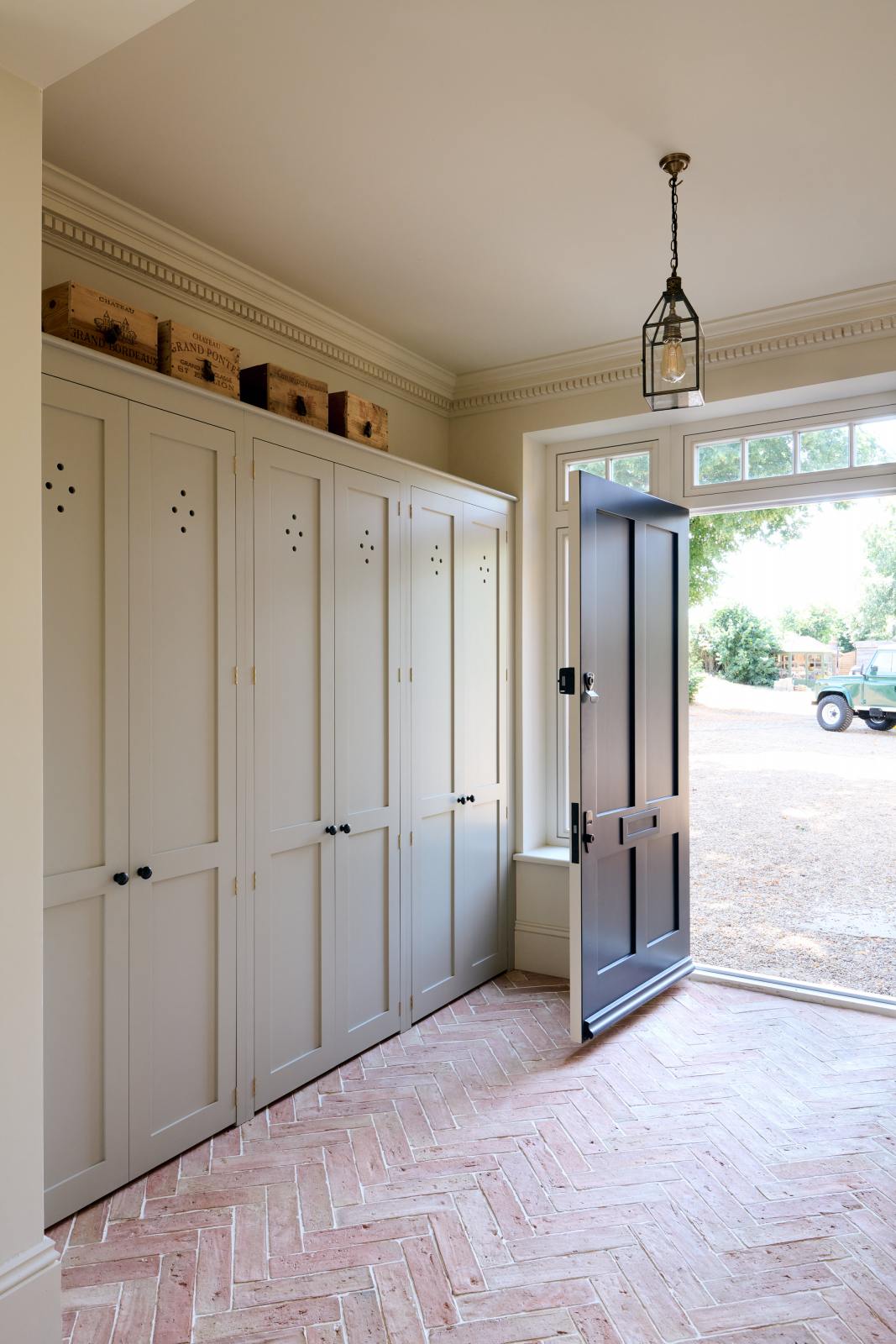 An oblique view of a modest foyer features bespoke storage cupboards in a muted sage green, complemented by a pale terracotta parquet floor, vintage wooden storage boxes, and a metal and glass pendant light, which adds to the coastal aesthetic.