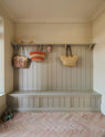A modest foyer featuring a wooden compartment bench, hooks, and a shelf painted in a muted sage green, complemented by the pale terracotta parquet floor, wicker bags, stray hats, and green and white striped slippers, adds to the coastal aesthetic.