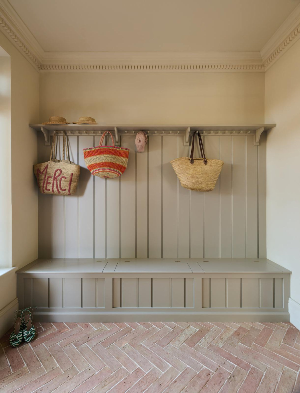 A modest foyer featuring a wooden compartment bench, hooks, and a shelf painted in a muted sage green, complemented by the pale terracotta parquet floor, wicker bags, stray hats, and green and white striped slippers, adds to the coastal aesthetic.