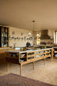 An angled view of a warm and chic Haberdasher’s kitchen features an L-shaped sink and cooker run, countertop cupboards, a kitchen island, a Carrara and copper mix worktop, and a double-fluted marble sink. Adorned with artisan ceramics, a deVOL gaselier light, Heirloom wall lights, and large vintage rugs, this kitchen creates a warm and inviting Spanish-inspired space in Yorkshire.