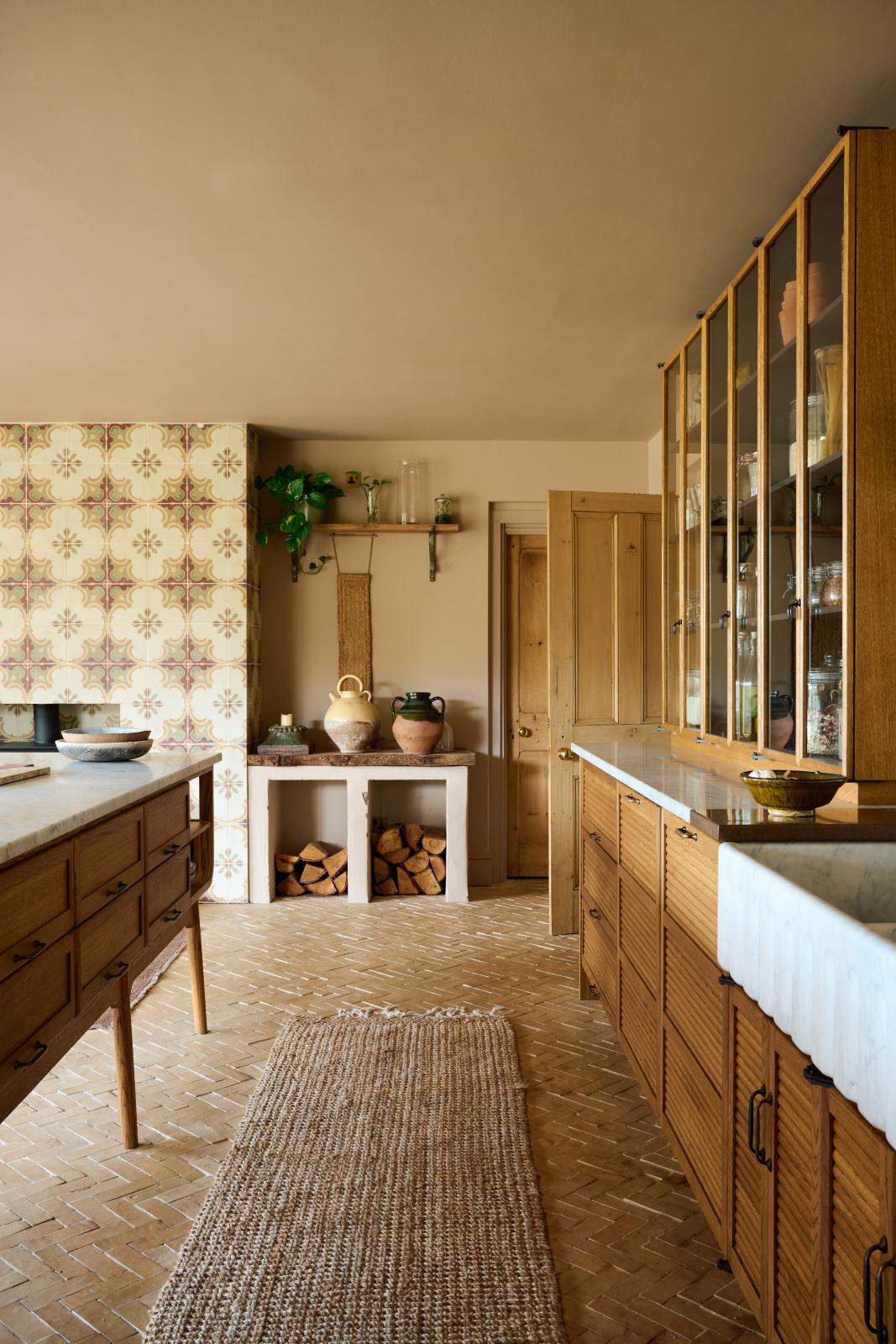 Nestled between a warm and chic Haberdasher’s kitchen features an L-shaped sink and cooker run, countertop cupboards, a kitchen island, a Carrara and copper mix worktop, and a double-fluted marble sink, is a firewood storage area. Adorned with artisan ceramics, a large vintage rug, and luscious green house plants, this kitchen creates a warm and inviting Spanish-inspired space in Yorkshire.