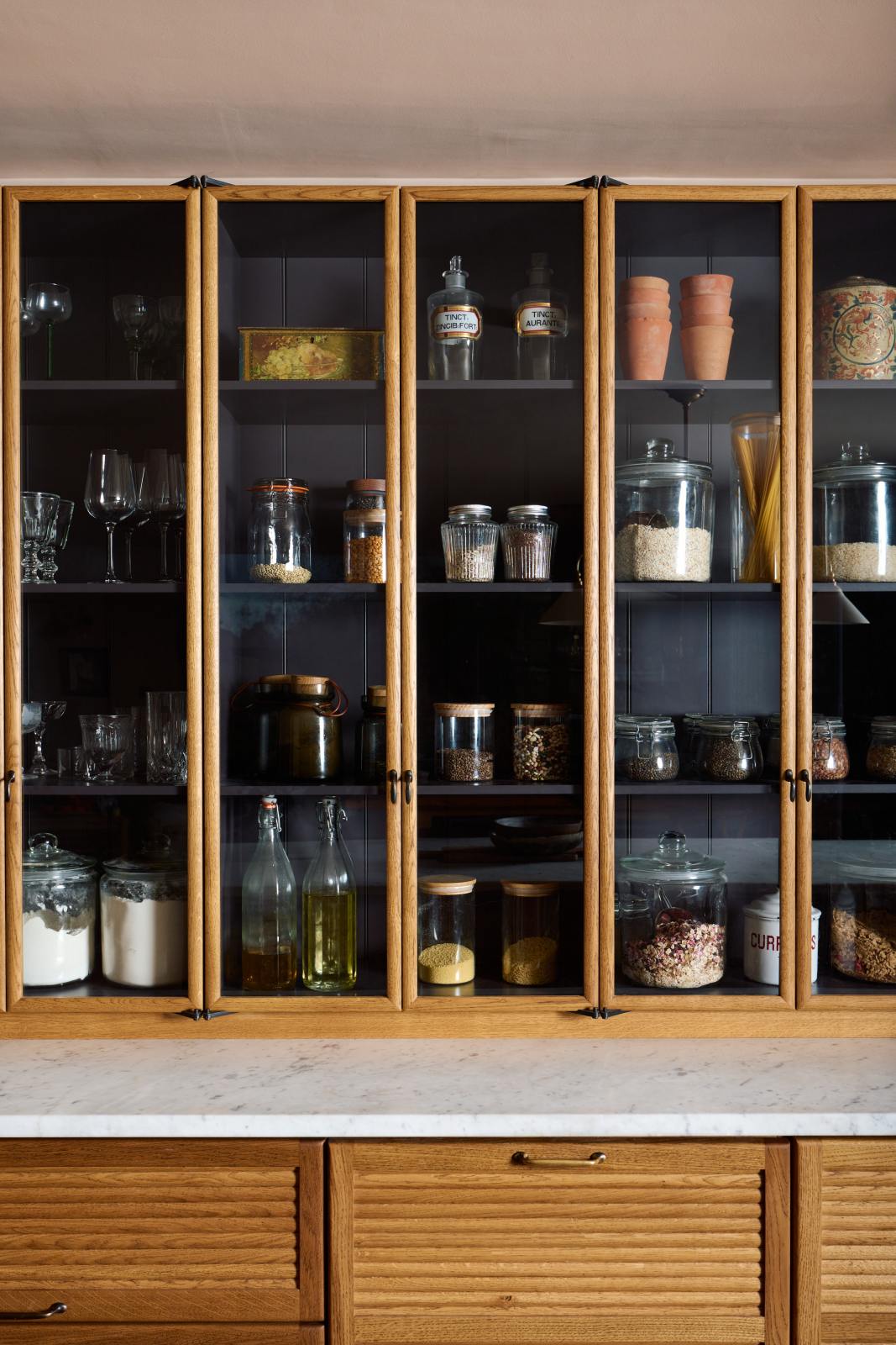 A close-up of a warm and chic Haberdasher’s kitchen features an L-shaped sink and cooker run, countertop cupboards, and a Carrara worktop. Adorned with artisan drinking glasses, dried ingredients in glass jars, clay pots, and vintage items, this kitchen creates a warm and inviting Spanish-inspired space in Yorkshire.