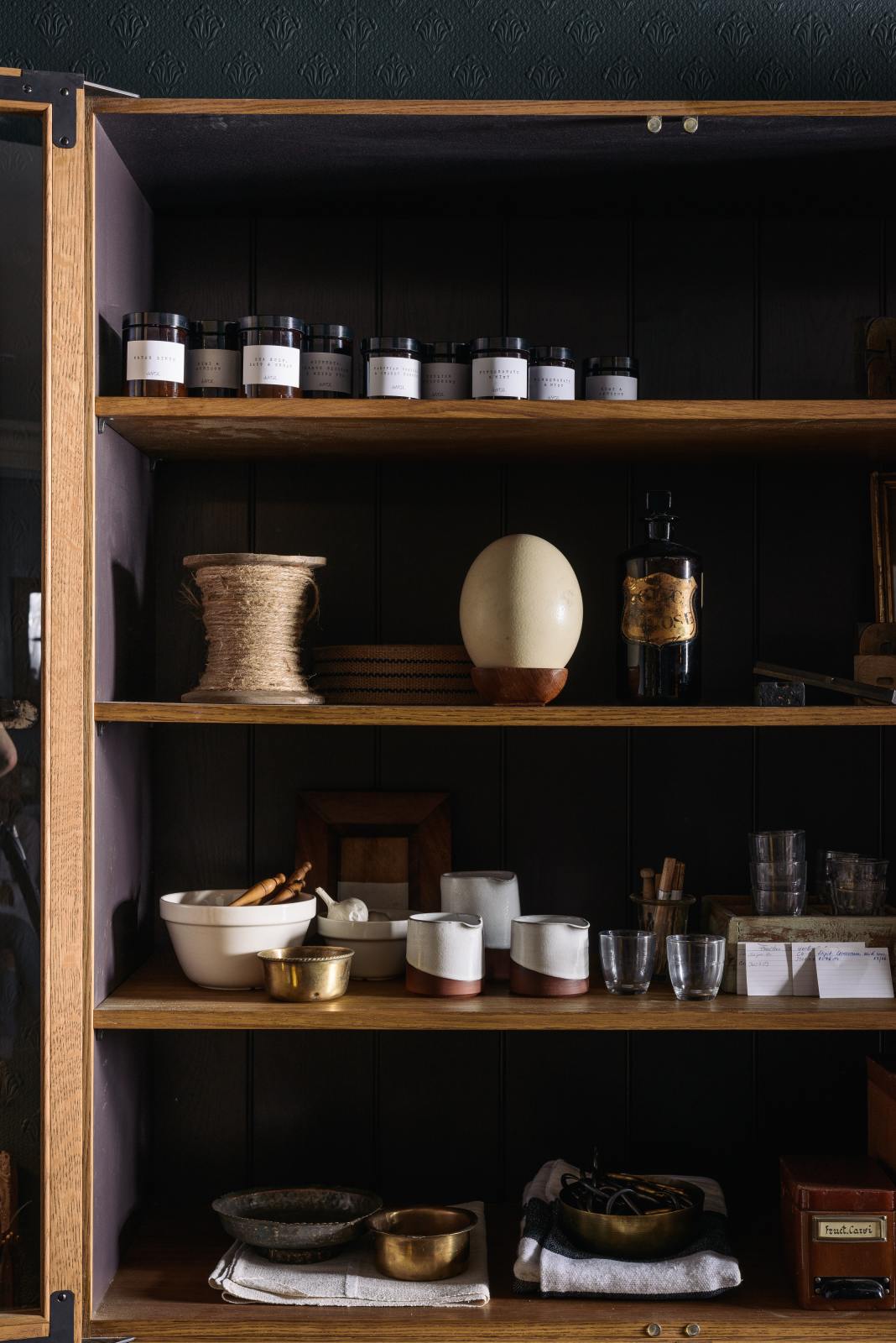 A closer look at a cool and elegant Haberdasher’s collection featuring a freestanding pantry with glazed doors. Adorned with vintage items, artisan ceramics, assorted containers, and candles, this space creates an atmospheric social bar.