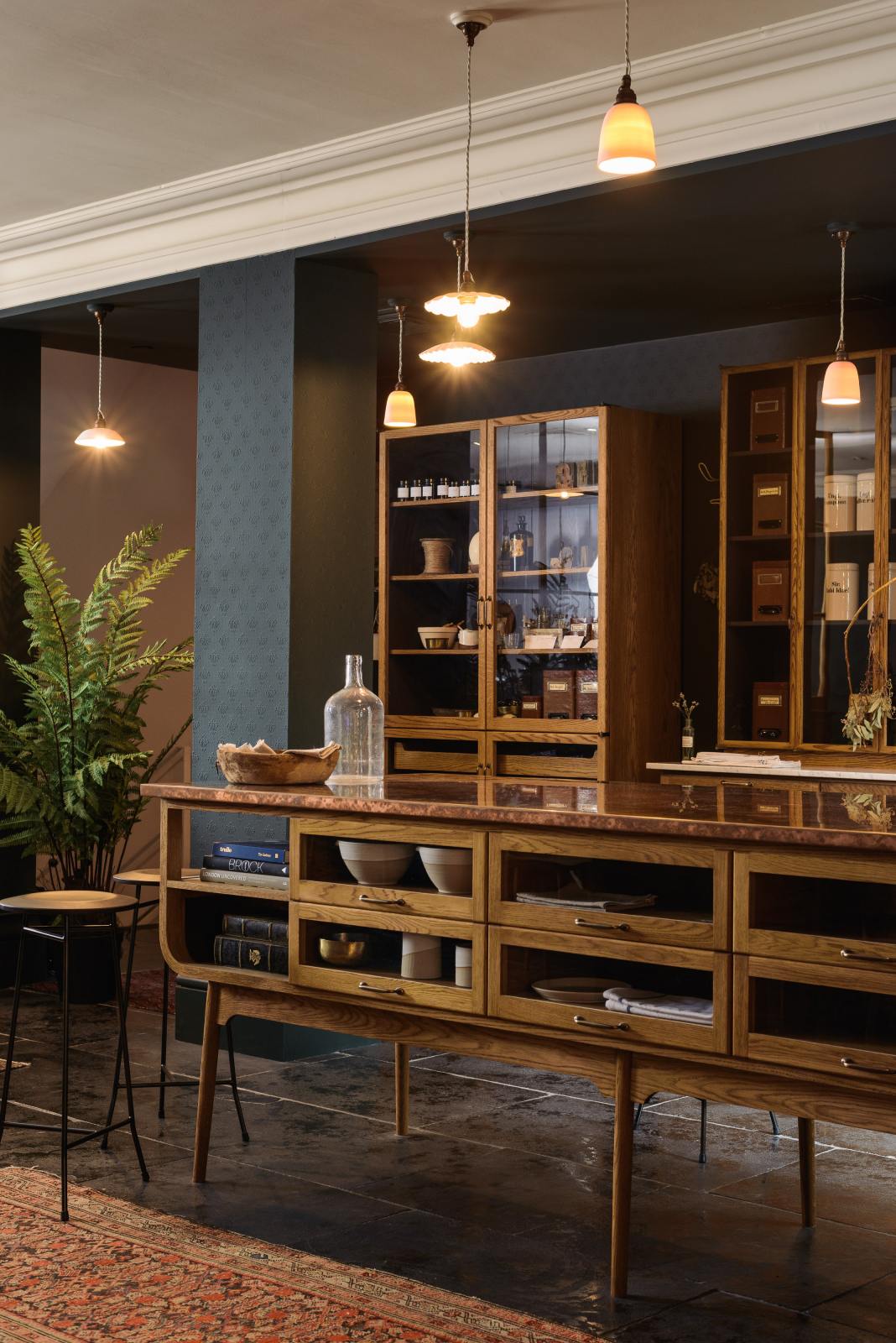 An oblique view of a cool and elegant Haberdasher’s collection features a large prep table with a copper worktop, a pantry run with tambour base cabinets, glazed countertop cabinets, and a freestanding pantry with glazed doors. Adorned with vintage furniture and items, artisan ceramics, the deVOL porcelain light collection, Creedy stools, assorted containers, and bottles, this space creates an atmospheric social bar.