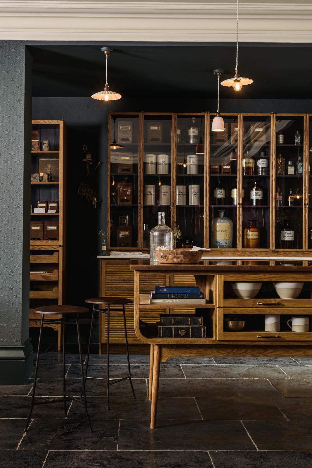 A cool and elegant Haberdasher’s collection features a large prep table with a copper worktop, a pantry run with tambour base cabinets, glazed countertop cabinets, and a freestanding pantry with glazed doors. Adorned with vintage furniture and items, artisan ceramics, the deVOL porcelain light collection, Creedy stools, assorted containers, and bottles, this space creates an atmospheric social bar.