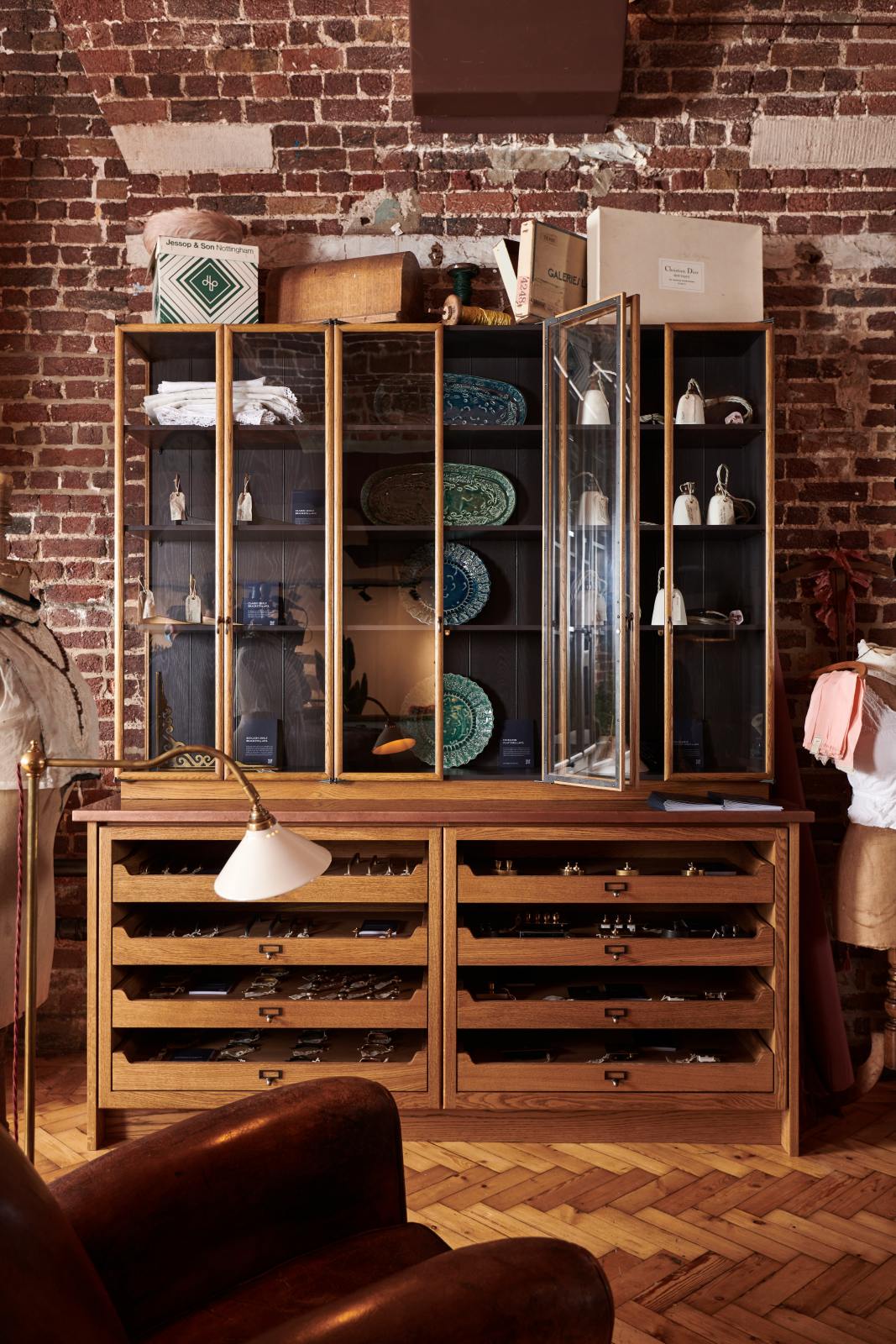 A closer look at the open base cabinets with internal drawers, countertop glazed cupboards, and a copper worktop as part of the handsome range of oak furniture and lighting from the deVOL Haberdasher’s collection. Adorned with the Grand tour reading lamp, vintage furniture and items, the Heirloom collection, and 1900s tailor tools, this space creates a beautiful homage to the English gentlemen’s haberdashery inspiration in a bricked wall showroom.