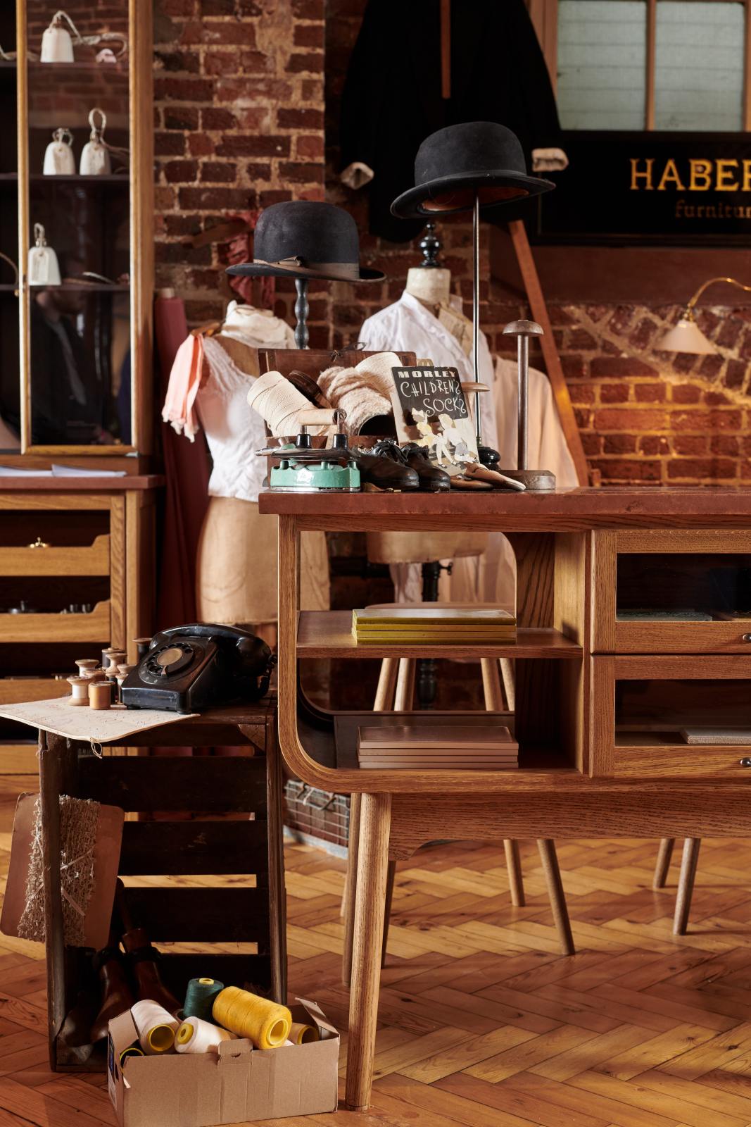 A closer look at a handsome range of oak furniture and lighting from the deVOL Haberdasher’s collection features a kitchen island, and countertop cupboards. Adorned with deVOL Heirloom accessories, Helen stools, vintage furniture and items, dressmaker mannequins, and 1900s tailor tools, this space creates a beautiful homage to the English gentlemen’s haberdashery inspiration in a bricked wall showroom.