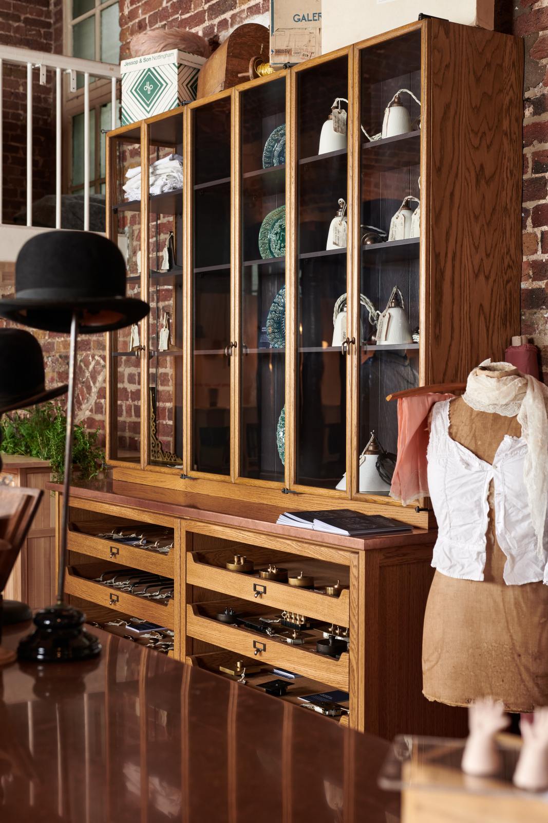 A close and oblique view of a handsome range of oak furniture and lighting from the deVOL Haberdasher’s collection features a kitchen island, a small run of open base cabinets, countertop cupboards, and a copper worktop. Adorned with deVOL Heirloom accessories, vintage furniture and items, dressmaker mannequins, and 1900s tailor tools, this space creates a beautiful homage to the English gentlemen’s haberdashery inspiration in a bricked wall showroom.