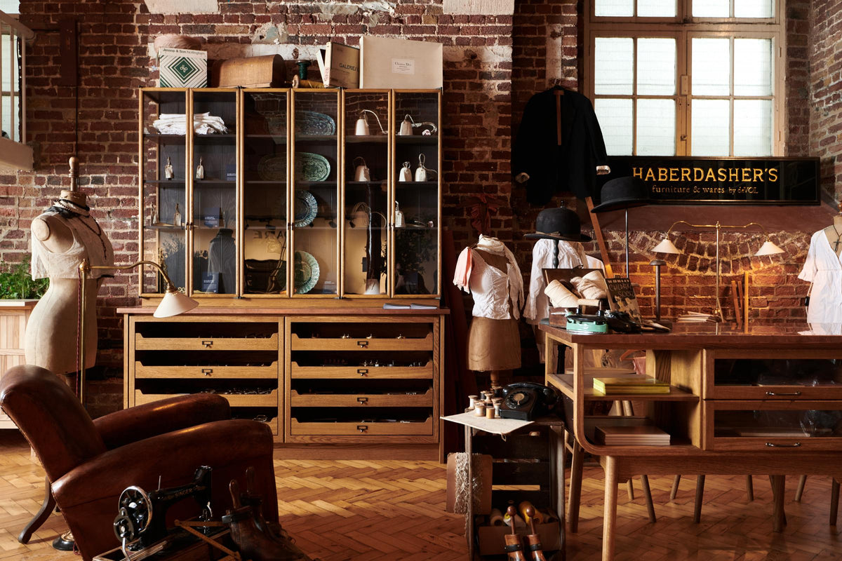 One side of a handsome range of oak furniture and lighting from the deVOL Haberdasher’s collection features a kitchen island, countertop cupboards, and copper countertop. Adorned with deVOL Heirloom accessories, Helen stools, vintage furniture and items, dressmaker mannequins, and 1900s tailor tools, this space creates a beautiful homage to the English gentlemen’s haberdashery inspiration in a bricked wall showroom.