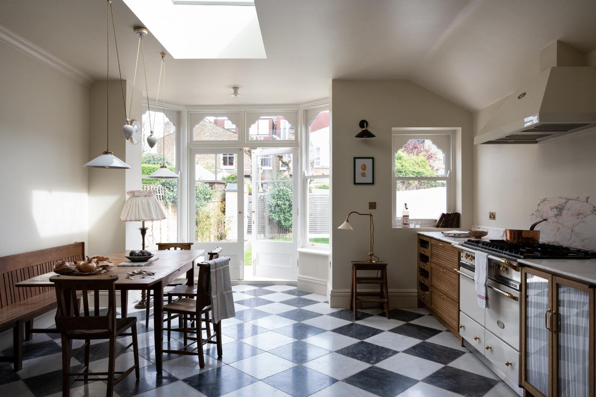 A grand and quaint Haberdasher’s kitchen features a sink and cooker run with a Paonazzo marble worktop and decorative vanity cut. Beautifully decorated with a white gas cooker with golden accents, a matching canopy, a deVOL grand tour desk lamp, vintage furniture, beloved chapel chairs, a skylight, and a cloth lampshade, this kitchen creates a humble and functional space with a touch of elegance.