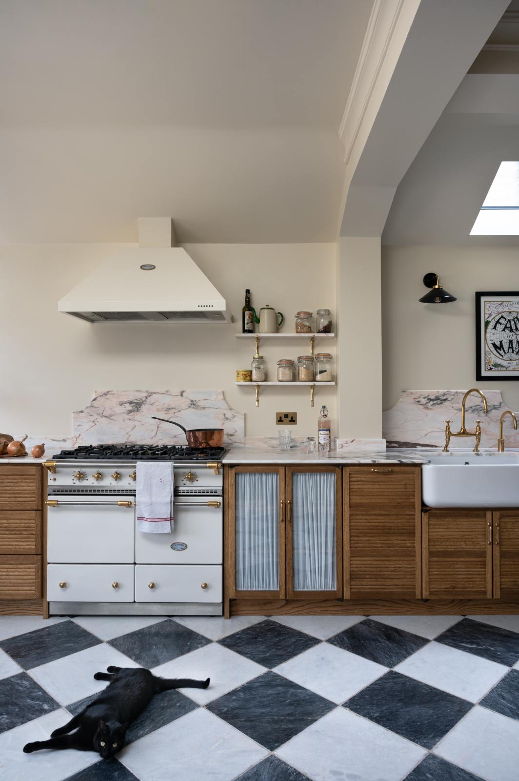 A grand and quaint Haberdasher’s kitchen features a sink and cooker run with a Paonazzo marble worktop and decorative vanity cut, a single Belfast sink, and an aged brass Ionian tap. Beautifully decorated with a white gas cooker with golden accents, a matching canopy, a deVOL grand tour desk lamp, vintage furniture, and a cute black cat lying on the chequerboard flooring, this kitchen creates a humble and functional space with a touch of elegance.