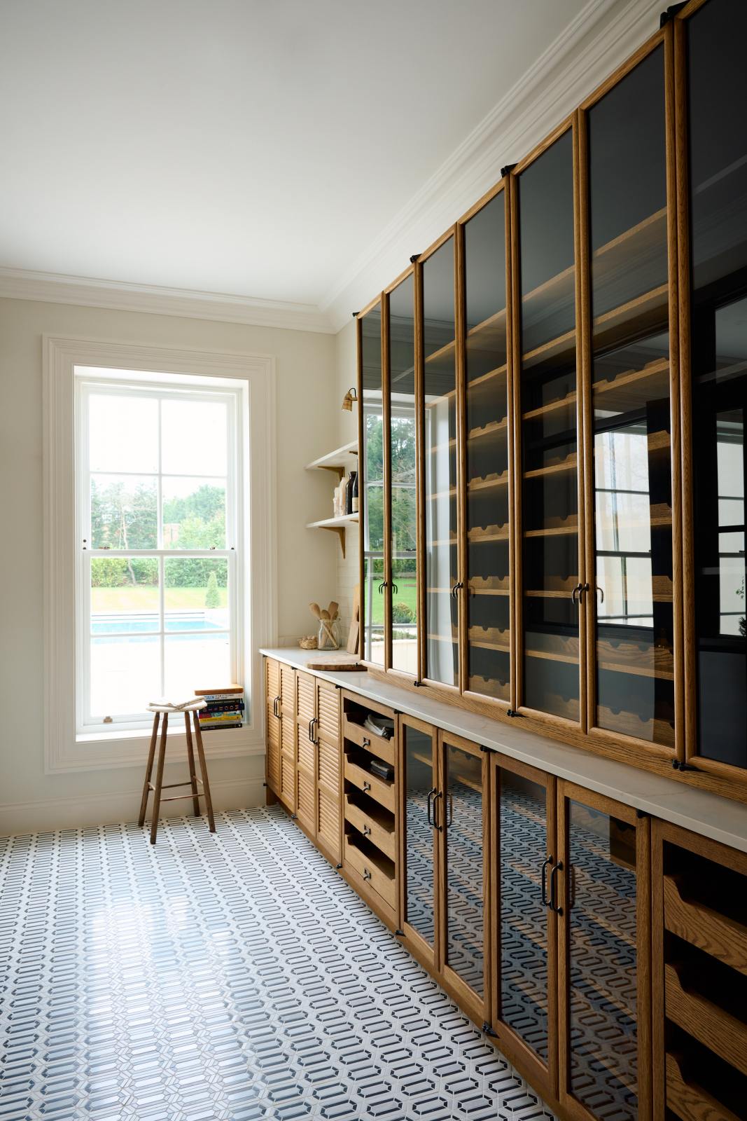 An oblique view of a smart and functional Haberdasher’s pantry features a long pantry run with glazed, open cupboards with internal drawers and tambour doors, a Carrara marble worktop and shelves, and cream splashback tiles. Beautifully decorated with eye-catching diamond mosaic flooring, vintage items, books, and tea towels, this pantry creates a warm and neat storage space.
