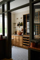 Past the olive green glass partition is a smart and functional Haberdasher’s pantry features a long pantry run with glazed, open cupboards with internal drawers and tambour doors, a Carrara marble worktop and shelves, and cream splashback tiles. Beautifully decorated with eye-catching diamond mosaic flooring, vintage items, cloves of garlic, onions, loaves of bread, and classic shelf brackets, this pantry creates a warm and neat storage space.