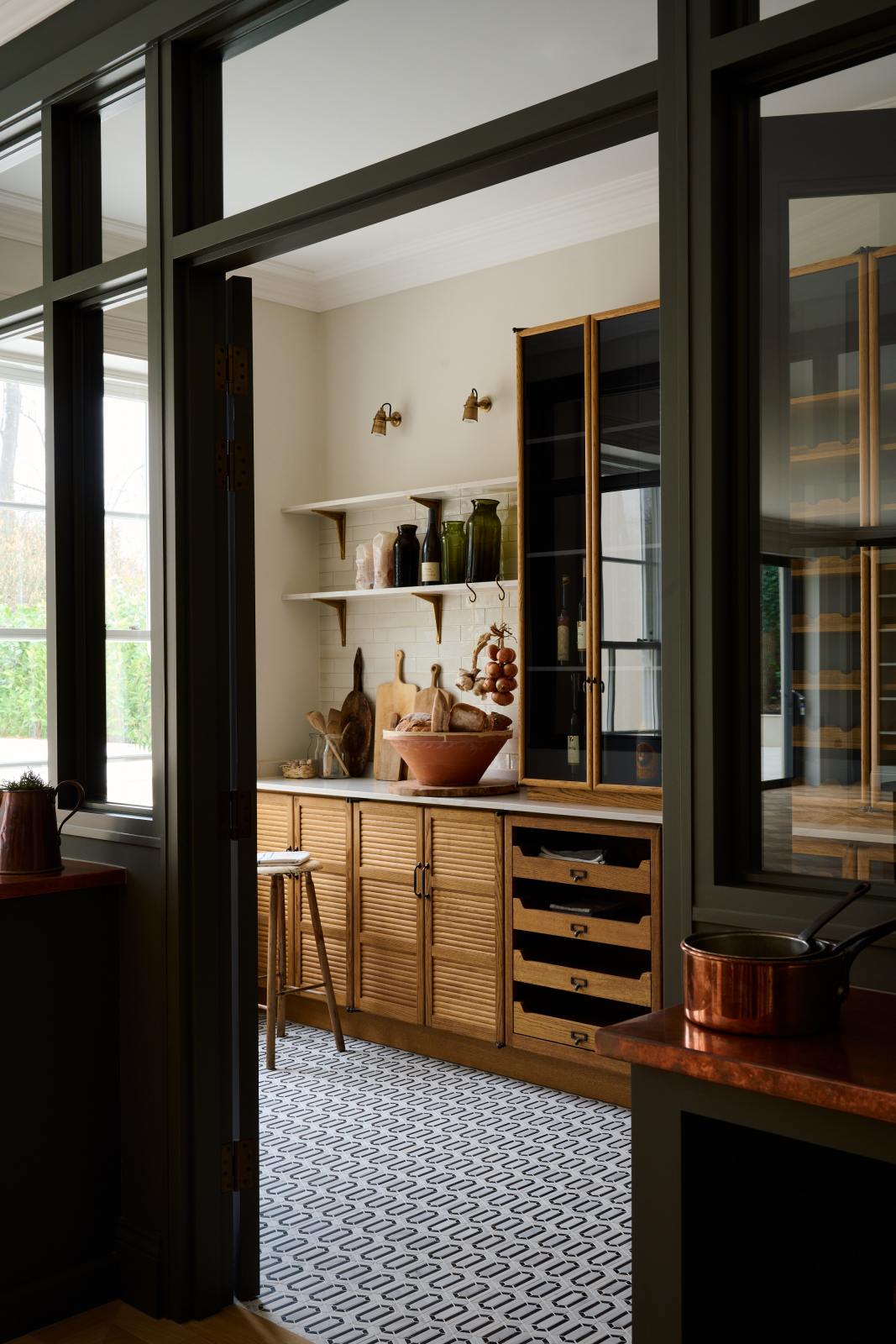 Past the olive green glass partition is a smart and functional Haberdasher’s pantry features a long pantry run with glazed, open cupboards with internal drawers and tambour doors, a Carrara marble worktop and shelves, and cream splashback tiles. Beautifully decorated with eye-catching diamond mosaic flooring, vintage items, cloves of garlic, onions, loaves of bread, and classic shelf brackets, this pantry creates a warm and neat storage space.