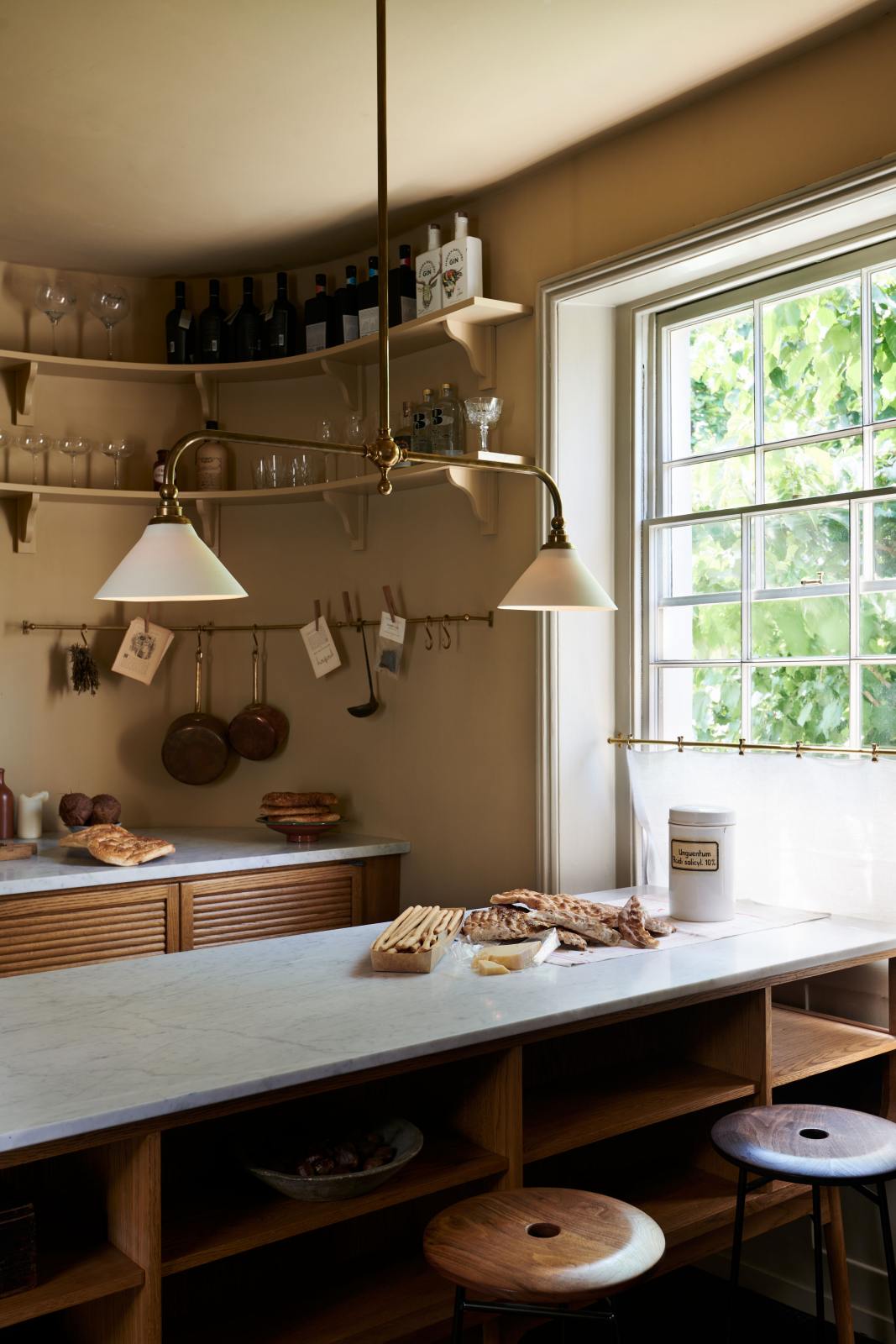 An oblique view of a cosy and sophisticated Haberdasher’s kitchen features a cooker run with a Carrara marble worktop, a small prep table, bespoke curved shelves, and muted yellow walls. Beautifully decorated with Creedy stools, a deVOL Heirloom gaselier light, aged brass hanging rails, copper pans, focaccia bread, vintage items, and artisan ceramics, this kitchen creates a bright and warm space.