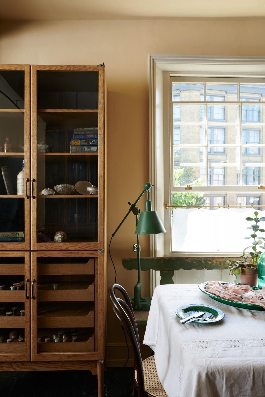 A cosy and sophisticated Haberdasher’s kitchen features a freestanding pantry with glazed doors. Beautifully decorated with Bentwood café chairs, vintage furniture and items, books, luscious green potted plants, aged brass café curtain rails, and artisan ceramics, this kitchen creates a bright and warm space.