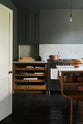 A cosy and sophisticated Haberdasher’s kitchen features a cooker run with a Carrara marble worktop, a small prep table, bespoke curved shelves, and dark charcoal walls. Beautifully decorated with stools, aged brass hanging rails, copper pans, fresh bread, vintage items, and artisan ceramics, this kitchen creates a cool yet warm and inviting space.