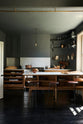 A cosy and sophisticated Haberdasher’s kitchen features a cooker run with a Carrara marble worktop, a small prep table, bespoke curved shelves, and dark charcoal walls. Beautifully decorated with stools, aged brass hanging rails, copper pans, fresh bread, vintage items, and artisan ceramics, this kitchen creates a cool yet warm and inviting space.
